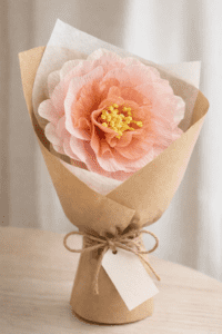 Grande pivoine en papier crépon rose, réalisée à la main et emballée dans un papier kraft façon fleuriste avec une étiquette “Bonne fête maman”.