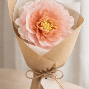 Grande pivoine en papier crépon rose, réalisée à la main et emballée dans un papier kraft façon fleuriste avec une étiquette “Bonne fête maman”.